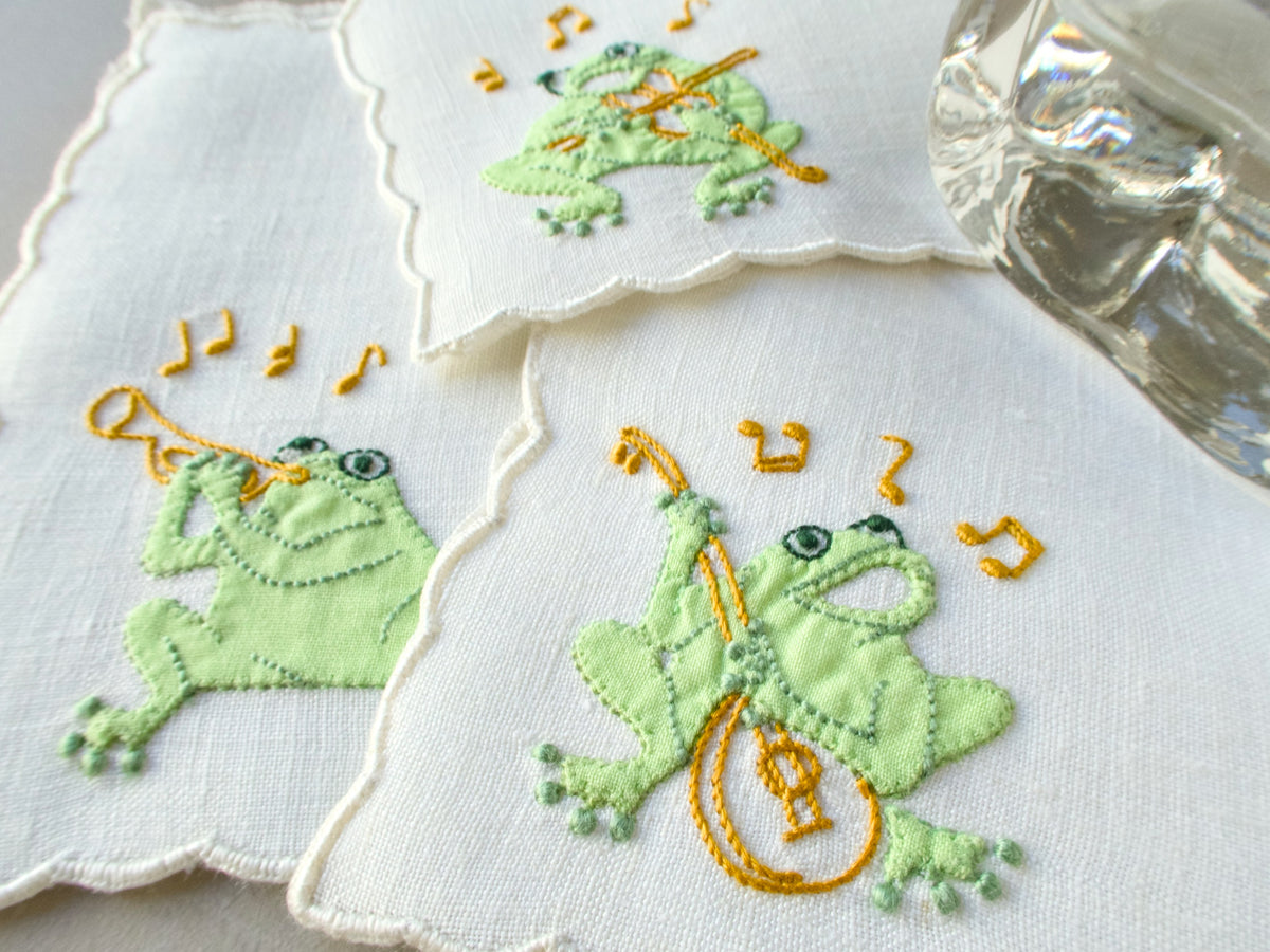 Close-up of vintage Madeira hand embroidered linen cocktail napkin with dimensional green frog playing a musical instrument, scalloped edges