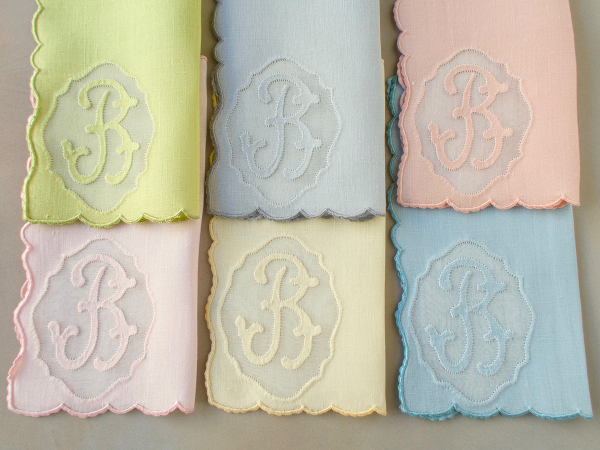 Set of six vintage Madeira cocktail napkins hand embroidered with cursive “B” monogram in organdy window, displayed in six pastel colors including blue, pink, yellow and green