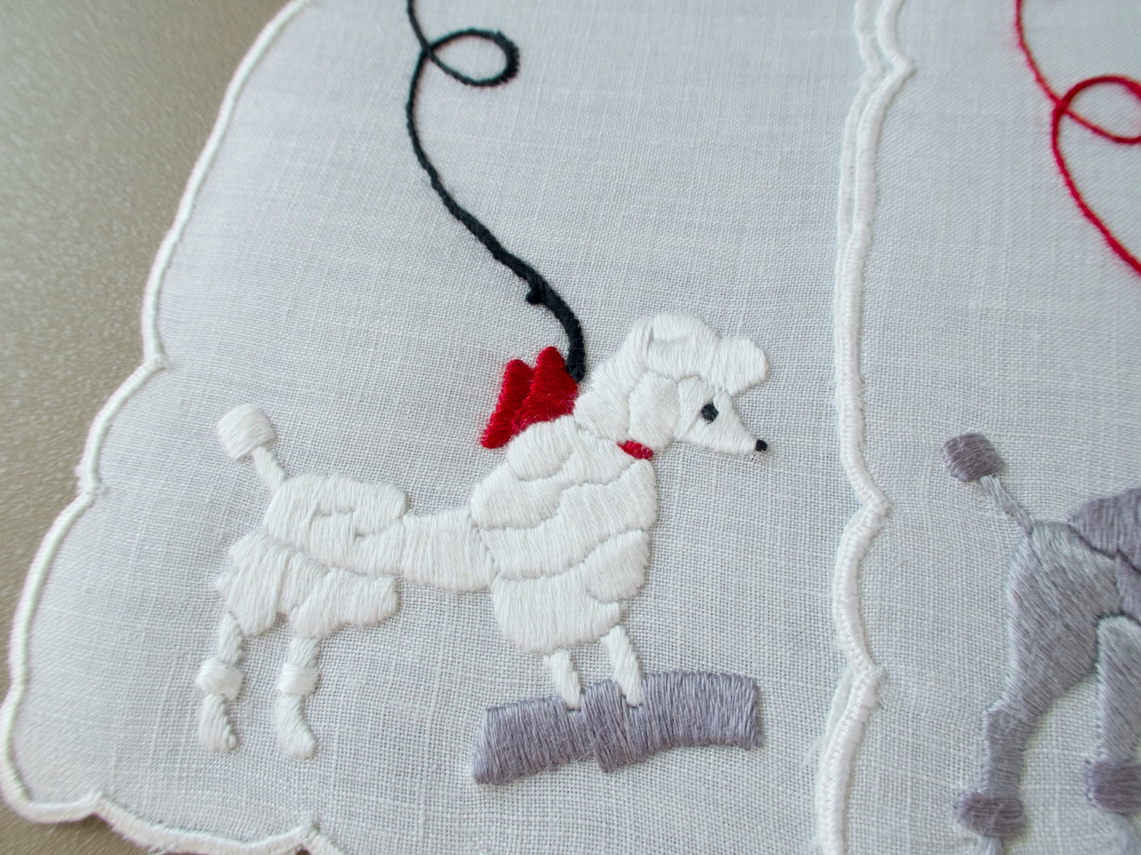 Close view of a white poodle in a red bow standing on a gray block, on a scalloped Madeira napkin