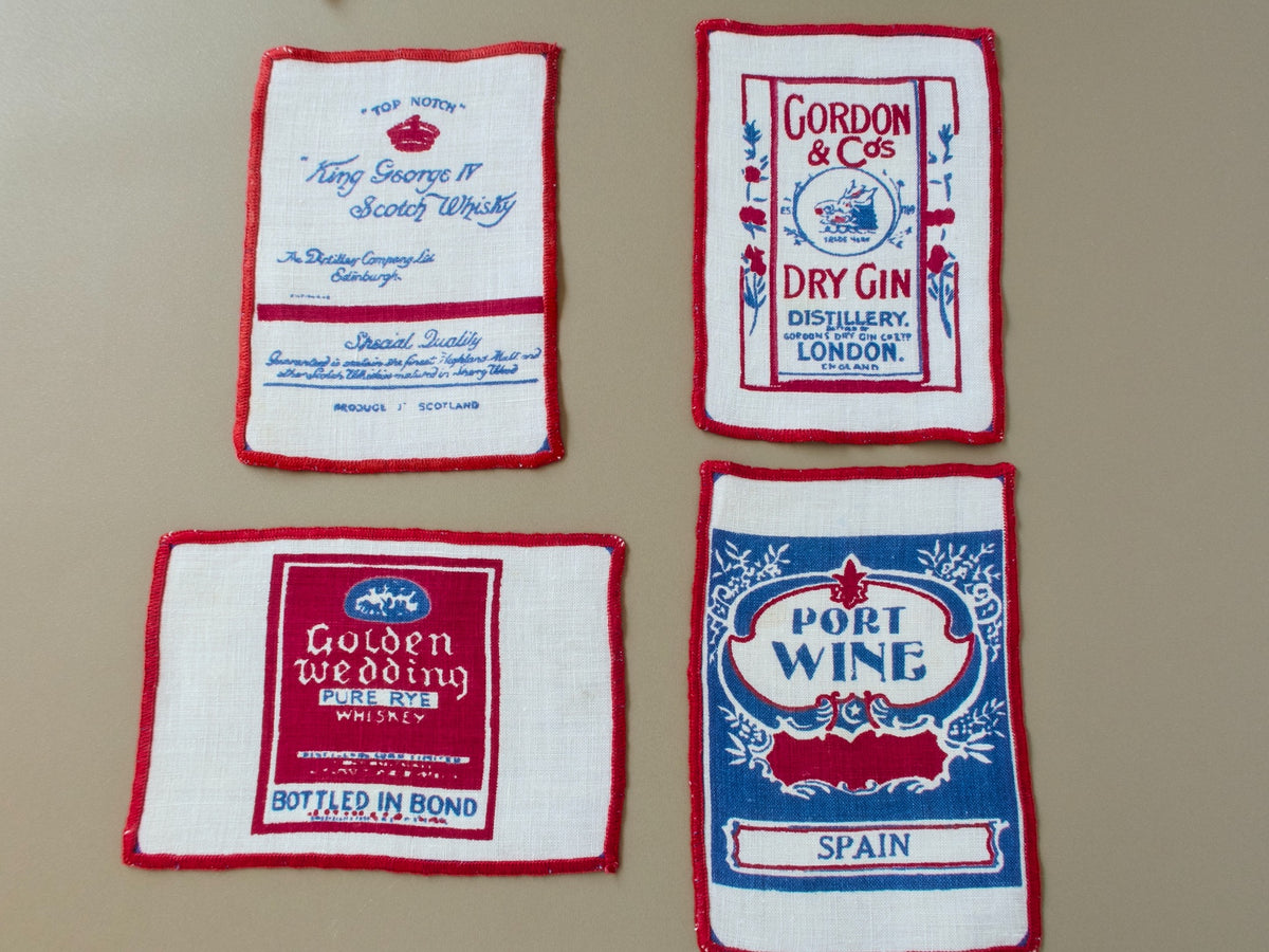 Four vintage linen cocktail napkins with red and blue printed liquor labels for Scotch, gin, port wine and rye whiskey.
