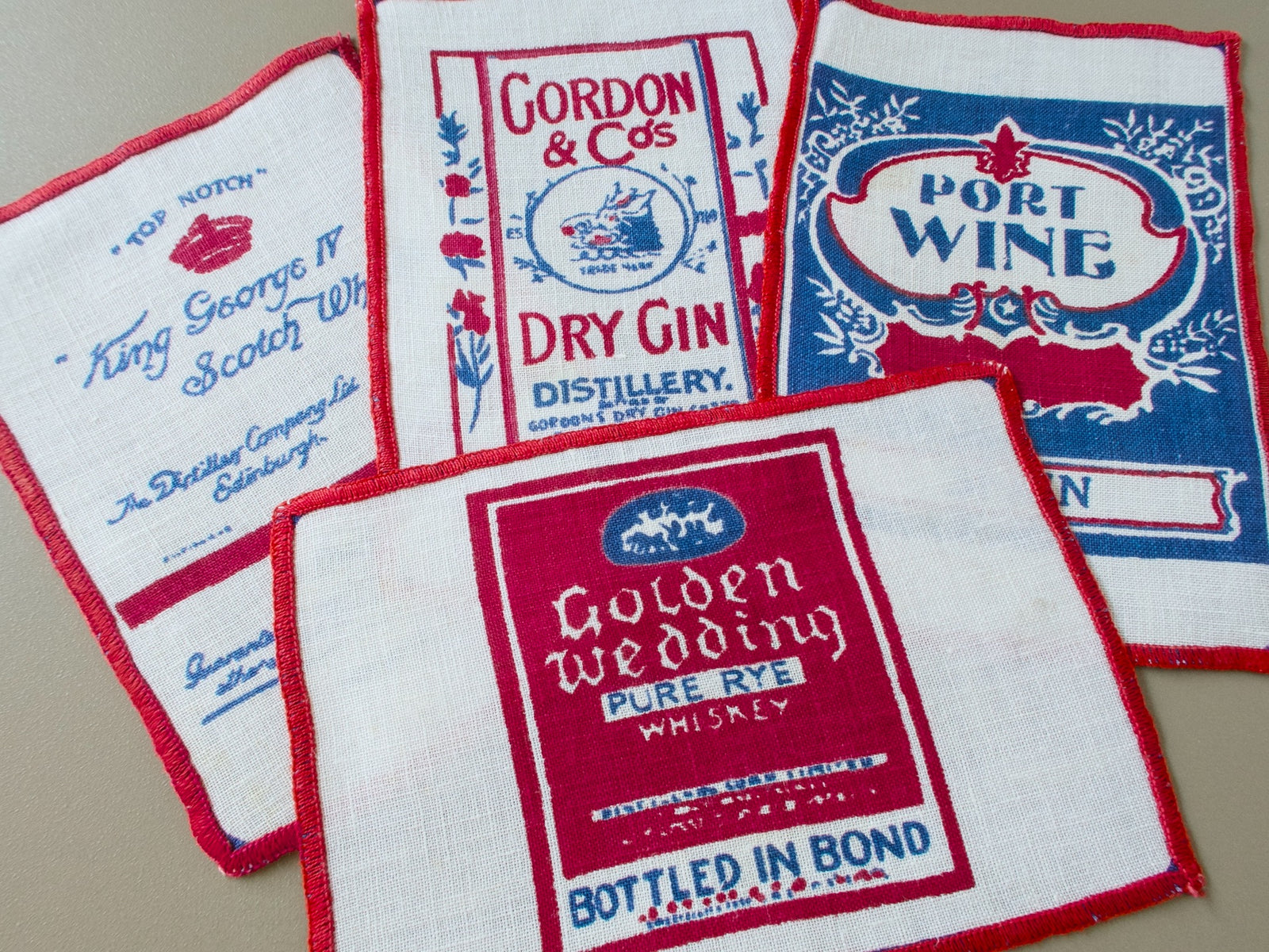 Four vintage linen cocktail napkins with red and blue printed liquor labels for Scotch, gin, port wine and rye whiskey.