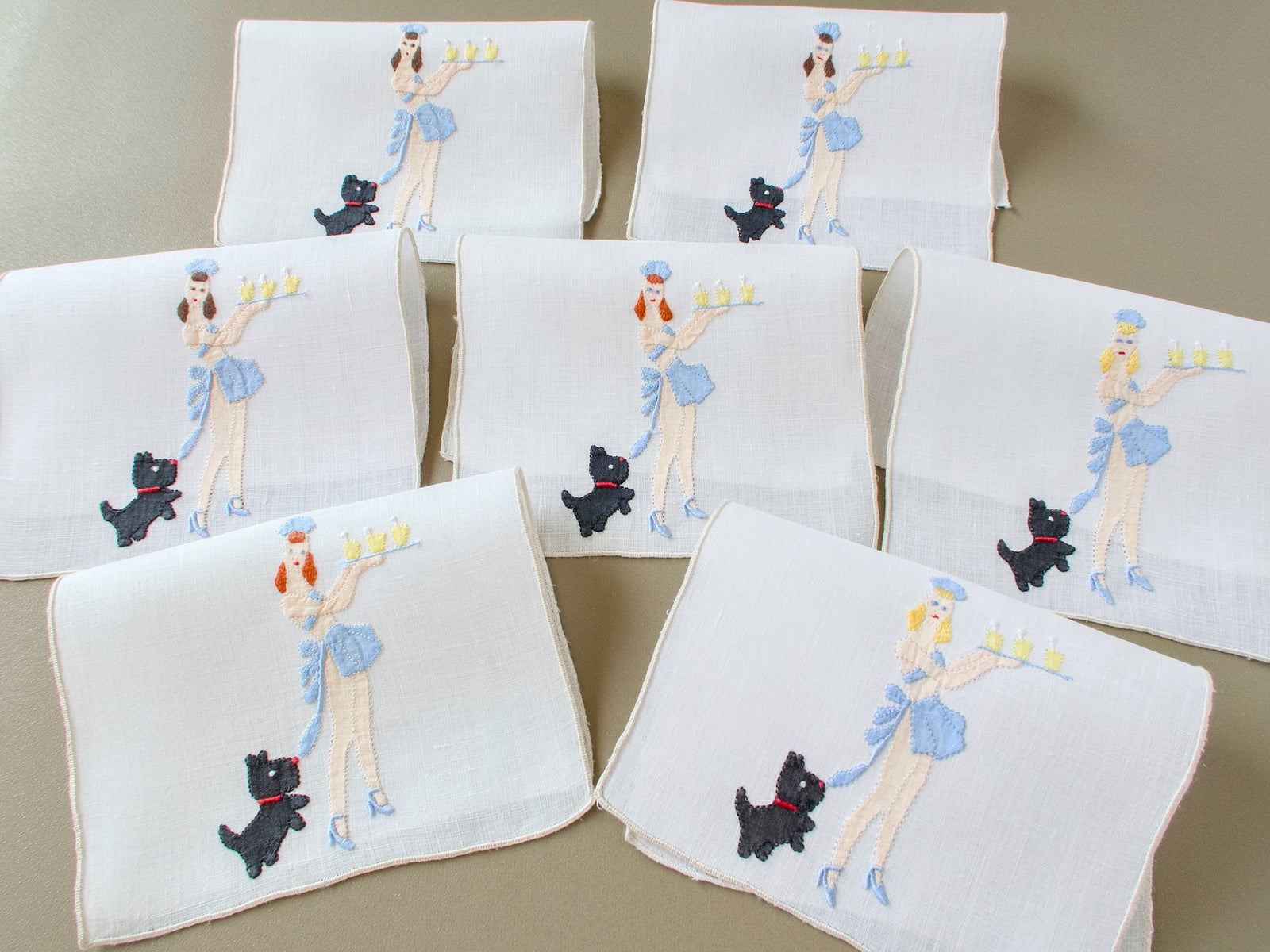A group of folded vintage Madeira linen cocktail napkins embroidered with a black dog tugging a waitress’s blue apron tie as she carries drinks.