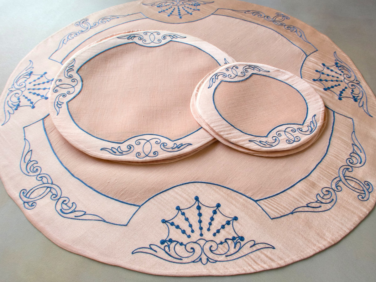 Full vintage French Cornely tea dessert set for 6 laid out, centerpiece with round placemat and coasters showing blue scrollwork embroidery on salmon linen​​​​​​​​​​​​​​​​