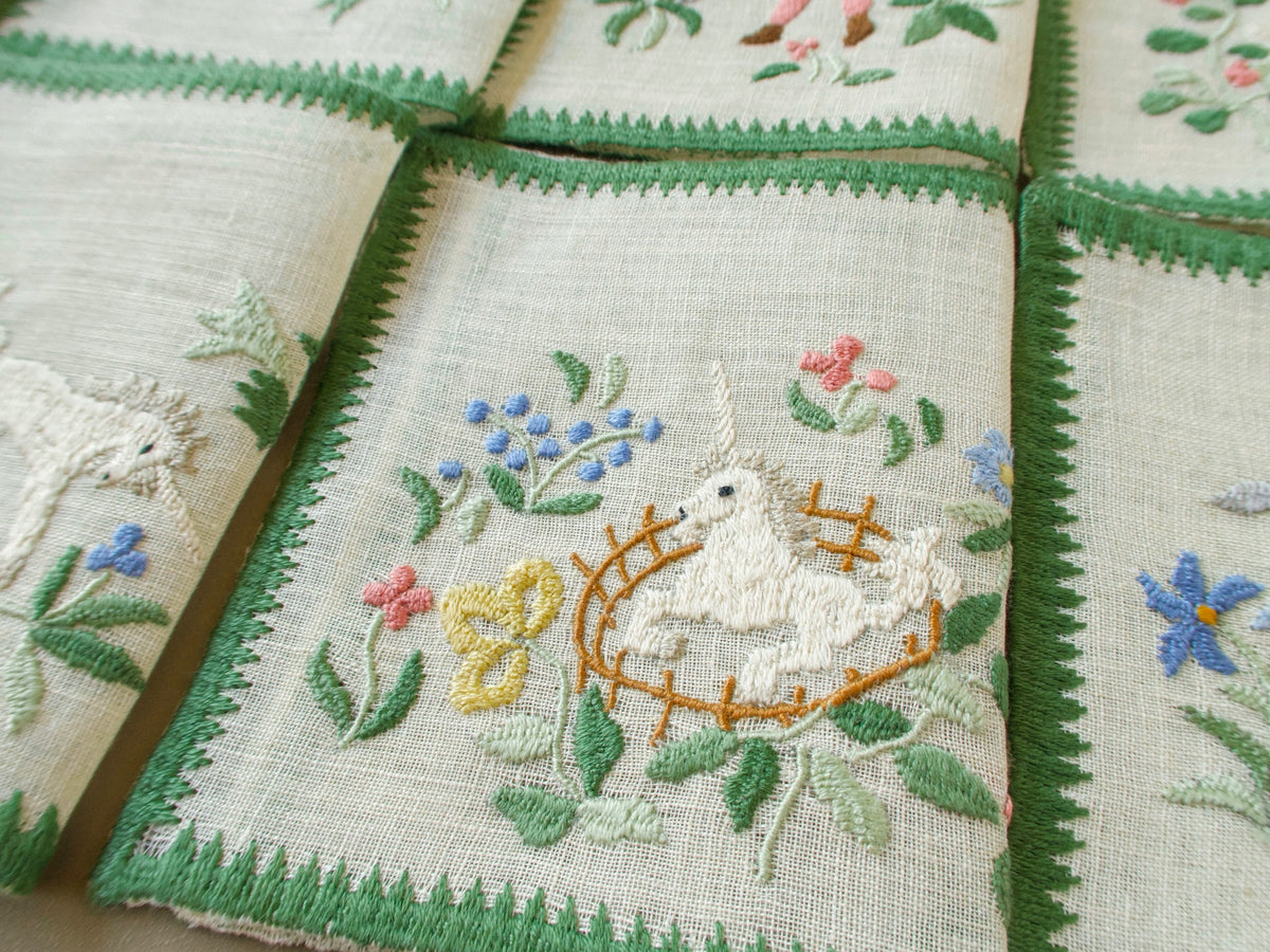 Vintage Baroness Rapisardi Hunt of the Unicorn cocktail napkin hand embroidered in punto stuoia with a unicorn inside a curved golden fence, surrounded by blue, pink and yellow flowers and edged in a green embroidered border.