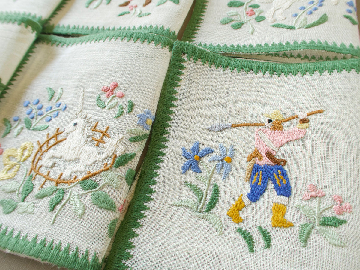 Close-up of two Baroness Rapisardi cocktail napkins showing the unicorn in a golden fence beside a hunter in a yellow hat, pink jacket, blue breeches and yellow boots aiming a long lance