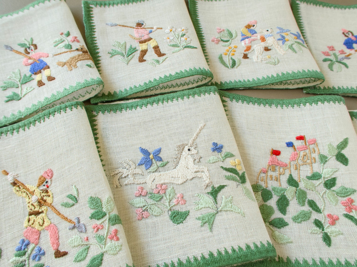 Two rows of Baroness Rapisardi cocktail napkins embroidered with spear-carrying hunters, a brown dog, a white hound, a leaping unicorn and a pink castle with red and blue flags
