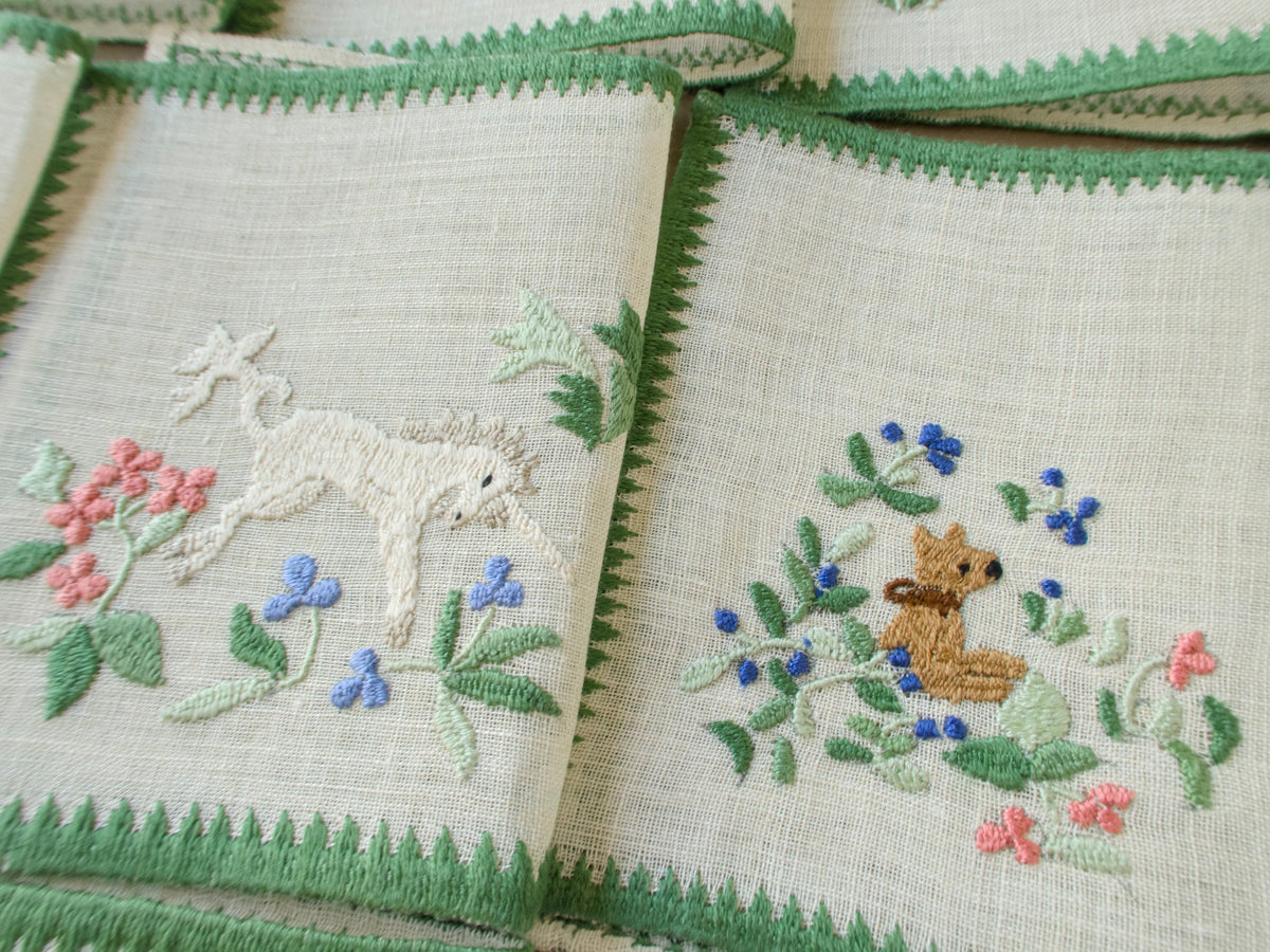 Close-up of two Baroness Rapisardi cocktail napkins showing a white unicorn with a gray mane and lowered head beside a small brown dog sitting among blue berries