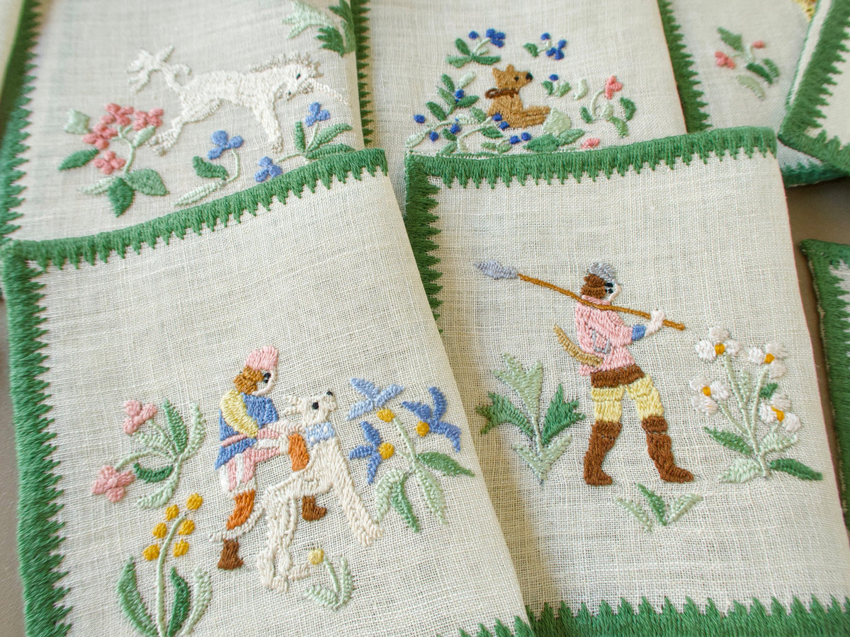 Four Baroness Rapisardi cocktail napkins showing a white unicorn, a small brown dog, a hunter with a white hound and a hunter carrying a spear over one shoulder, all among flowers