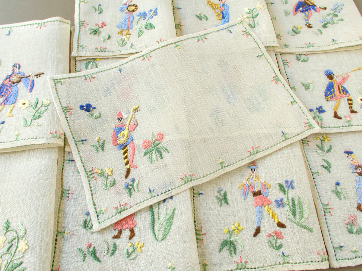 A vintage Baroness Rapisardi Medieval Musicians cocktail napkin opened flat to show the semi-sheer linen, with a lute player in a pink cap and striped hose embroidered at one end and other musician napkins folded beneath