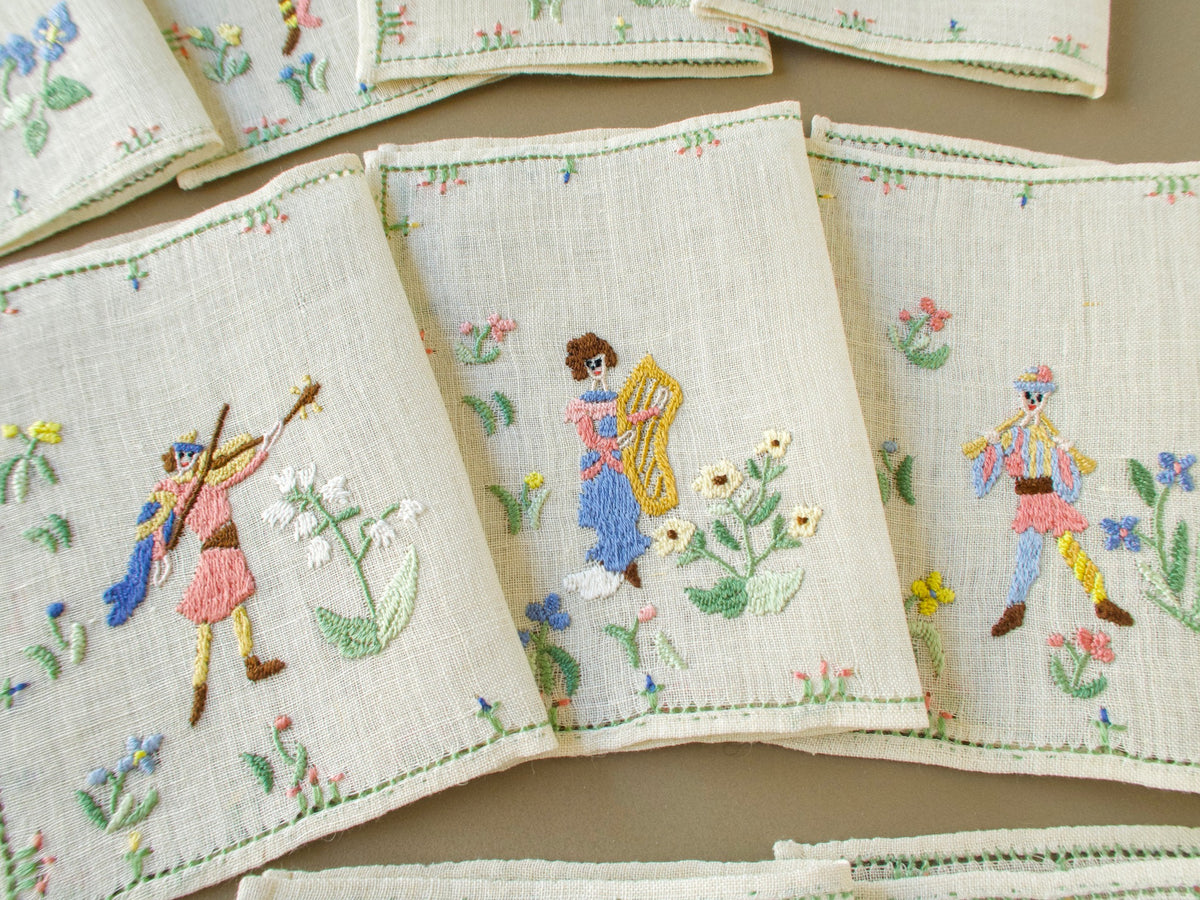 Three Baroness Rapisardi cocktail napkins showing a violinist in a pink tunic and blue cape, a harpist with brown hair holding a gold harp and a piper in a striped tunic, each among flowers