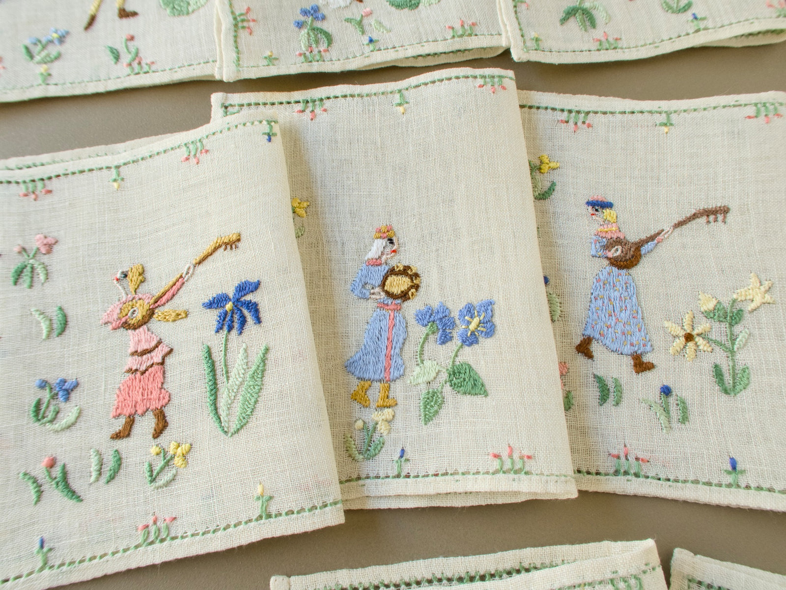 Three Baroness Rapisardi cocktail napkins showing a lute player in a pink tunic, a tambourine player in a blue robe and a lute player in a blue dotted dress, with iris, violets and yellow flowers