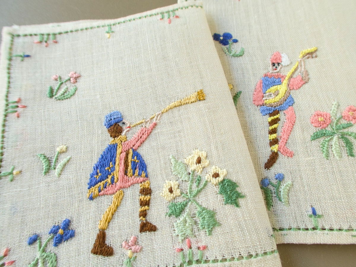 Close-up of two Baroness Rapisardi cocktail napkins showing a horn player in a gold trimmed blue cape beside a lute player in a pink and blue tunic and striped hose, with yellow and pink flowers