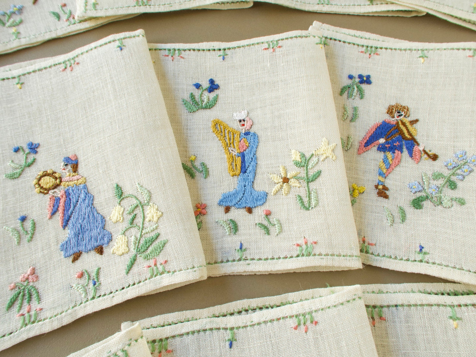 Three Baroness Rapisardi cocktail napkins showing a tambourine player in a blue robe, a harpist in a blue gown with a gold harp and a fiddler in a blue and pink cape, with green hemstitched borders