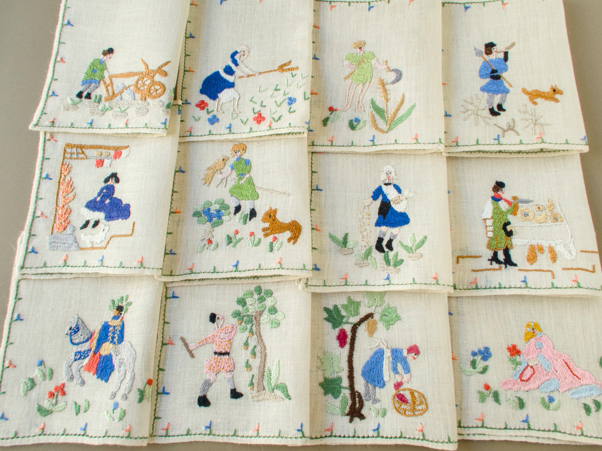 Set of 12 folded vintage Baroness Rapisardi Months of the Year cocktail napkins in three rows, each hand embroidered with a different seasonal scene from plowing and haying to the grape harvest and winter hunt, using the punto stuoia technique