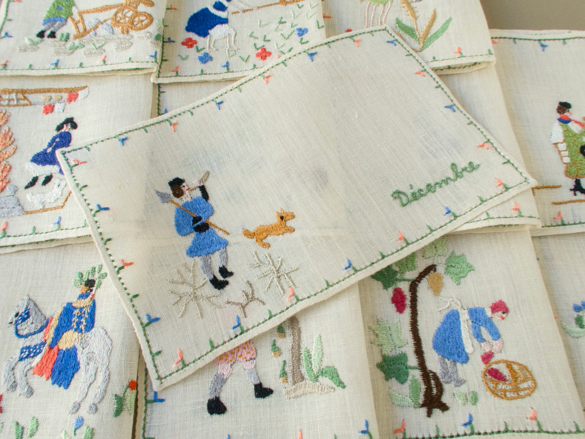 Baroness Rapisardi Décembre cocktail napkin opened flat to show a hunter in blue with a spear and horn, a small tan dog and frosted branches, with the month name in green script at right
