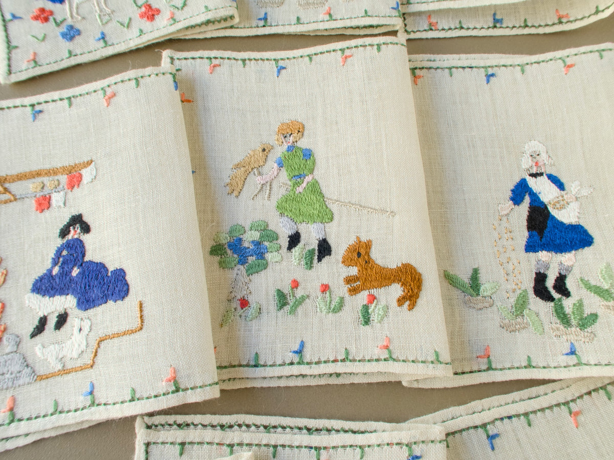Close-up of Baroness Rapisardi cocktail napkins showing a falconer with a bird and a leaping tan dog beside a sower in a blue tunic scattering seed and a figure seated by a hearth with a white dog
