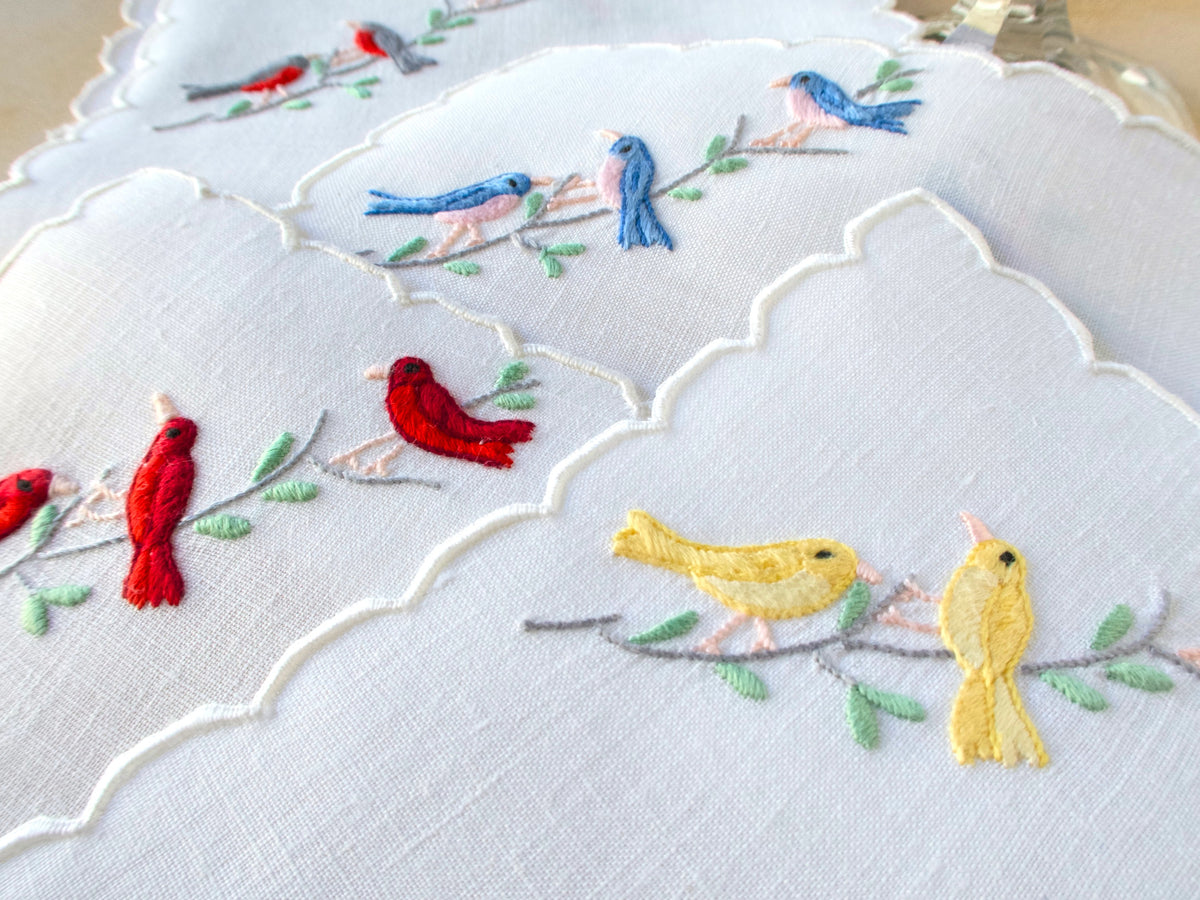 Close-up of colorful hand embroidered songbirds on white vintage Madeira linen cocktail napkins with scalloped edges.