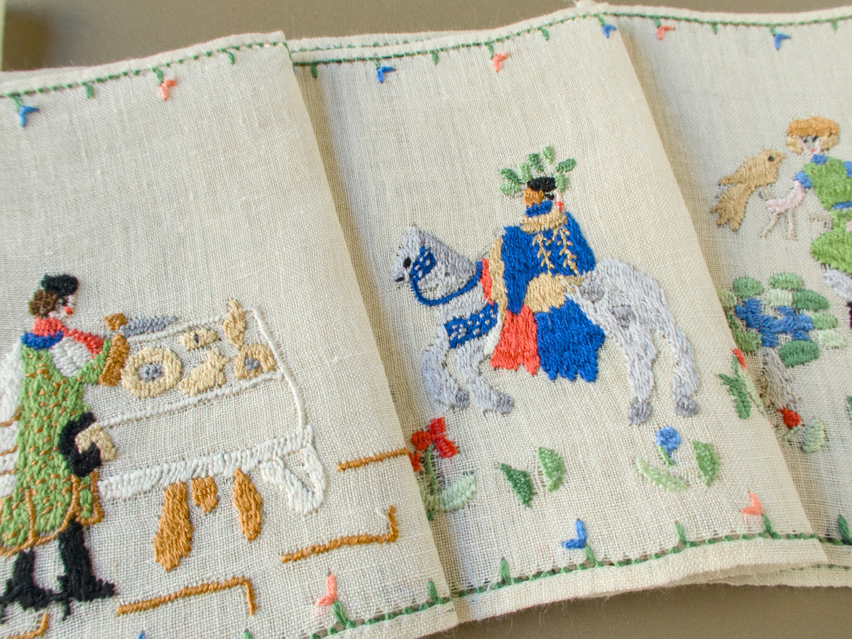 Close-up of Baroness Rapisardi cocktail napkins showing a figure in a green coat at a table set with round loaves and pastries beside a rider in a blue cloak on a gray horse
