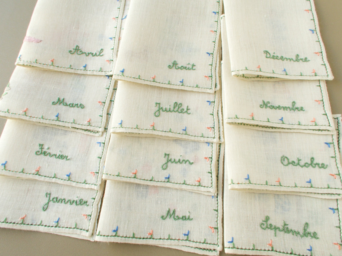 All 12 Baroness Rapisardi cocktail napkins folded to show the month names from Janvier to Décembre embroidered in green script above small pink and blue flower borders