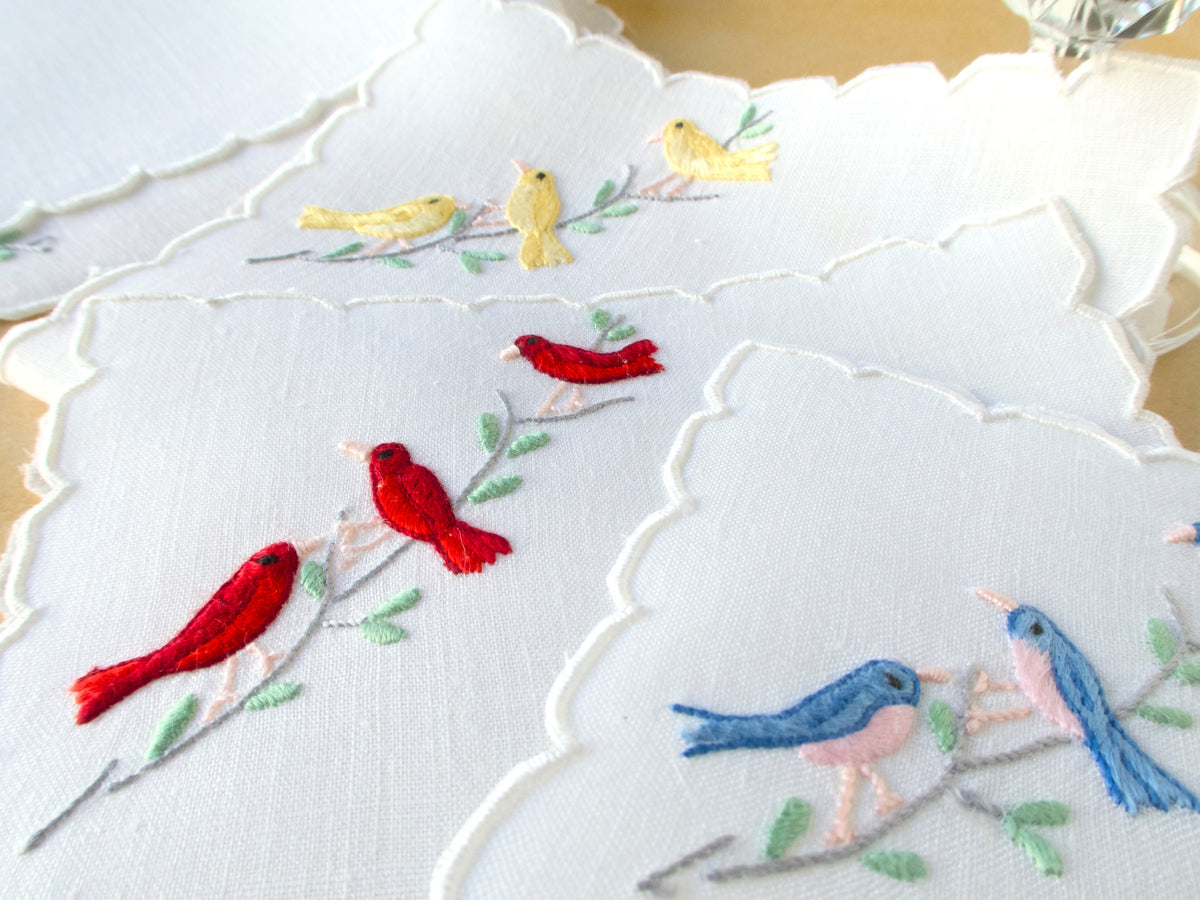 Close-up of colorful hand embroidered songbirds on white vintage Madeira linen cocktail napkins with scalloped edges.