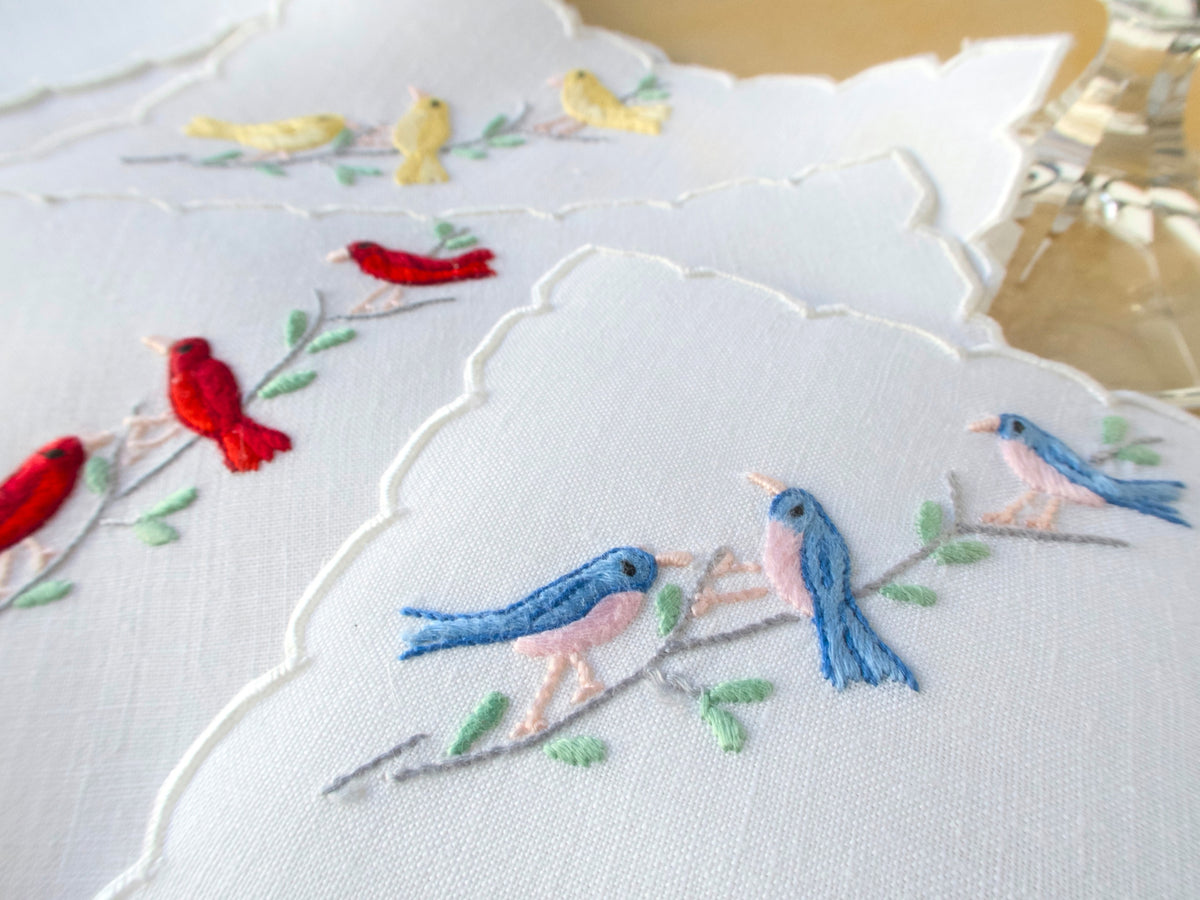 Close-up of colorful hand embroidered songbirds on white vintage Madeira linen cocktail napkins with scalloped edges.