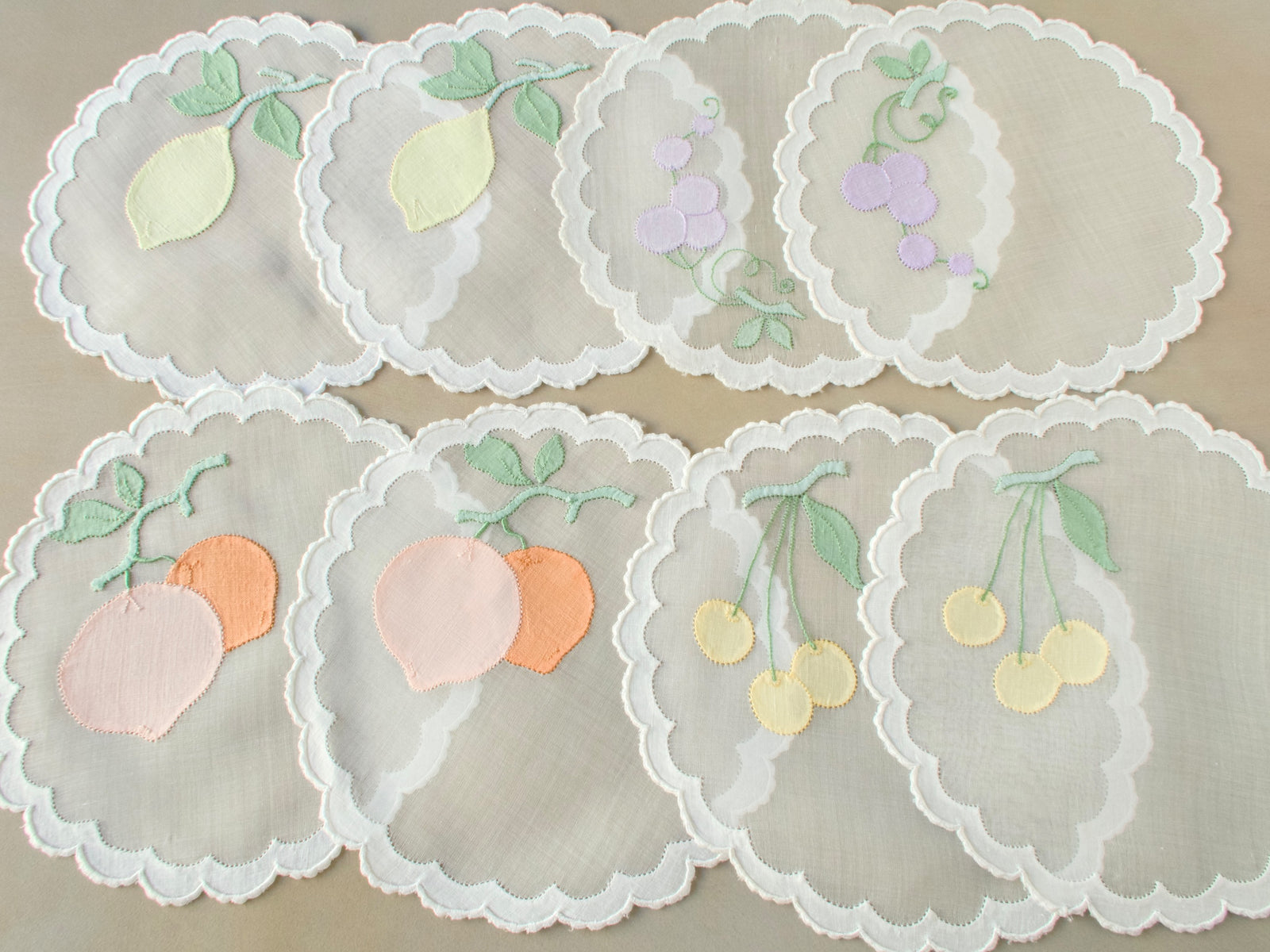 Overhead view of eight vintage Madeira organdy cocktail rounds hand embroidered with fruit appliqué in soft colors, arranged in a circle showing scalloped edging detail​​​​​​​​​​​​​​​​
