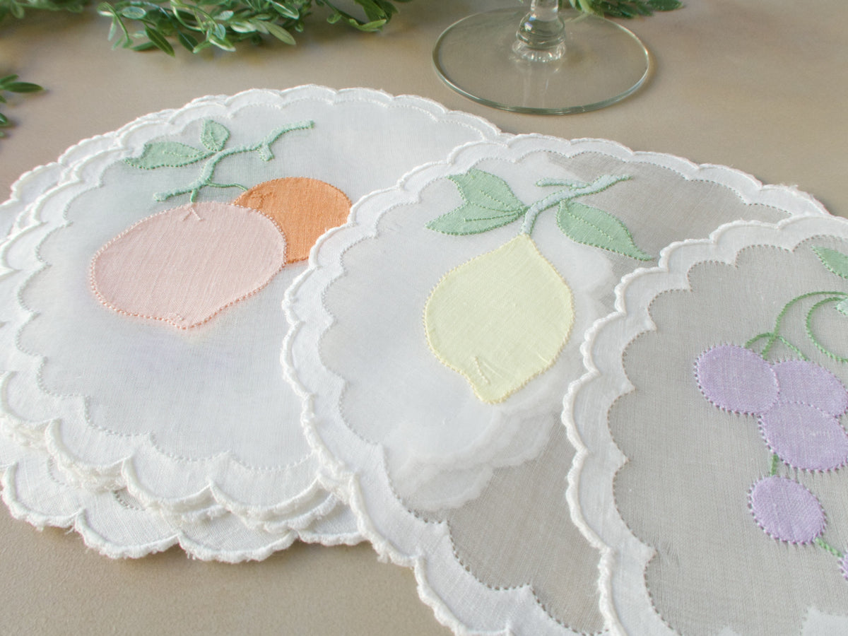 Vintage Madeira organdy cocktail rounds hand embroidered with soft-colored fruit appliqué and caseado bastido edging, fanned out on table with crystal stemware, set of 8