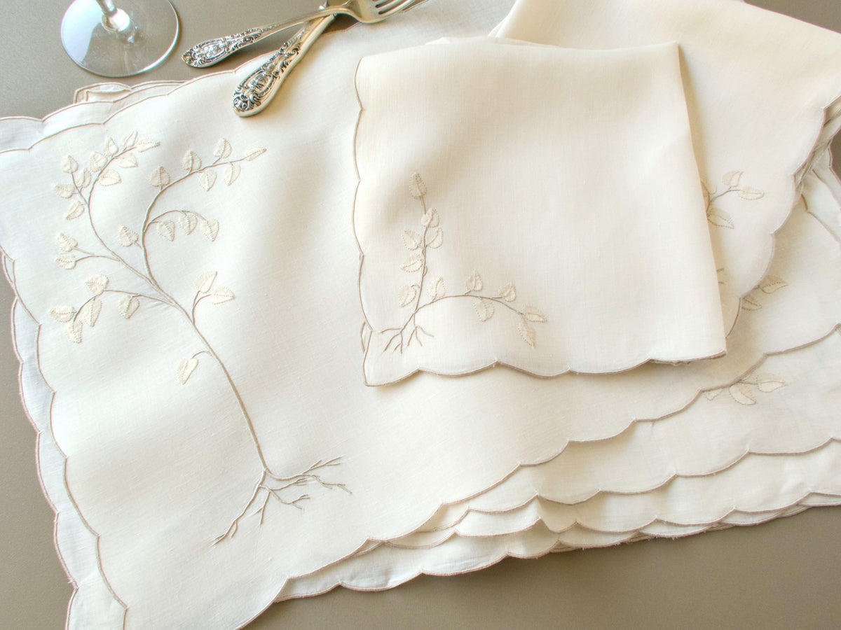 Stack of vintage Madeira linen placemats with scalloped edges and a matching napkin beside antique silver flatware
