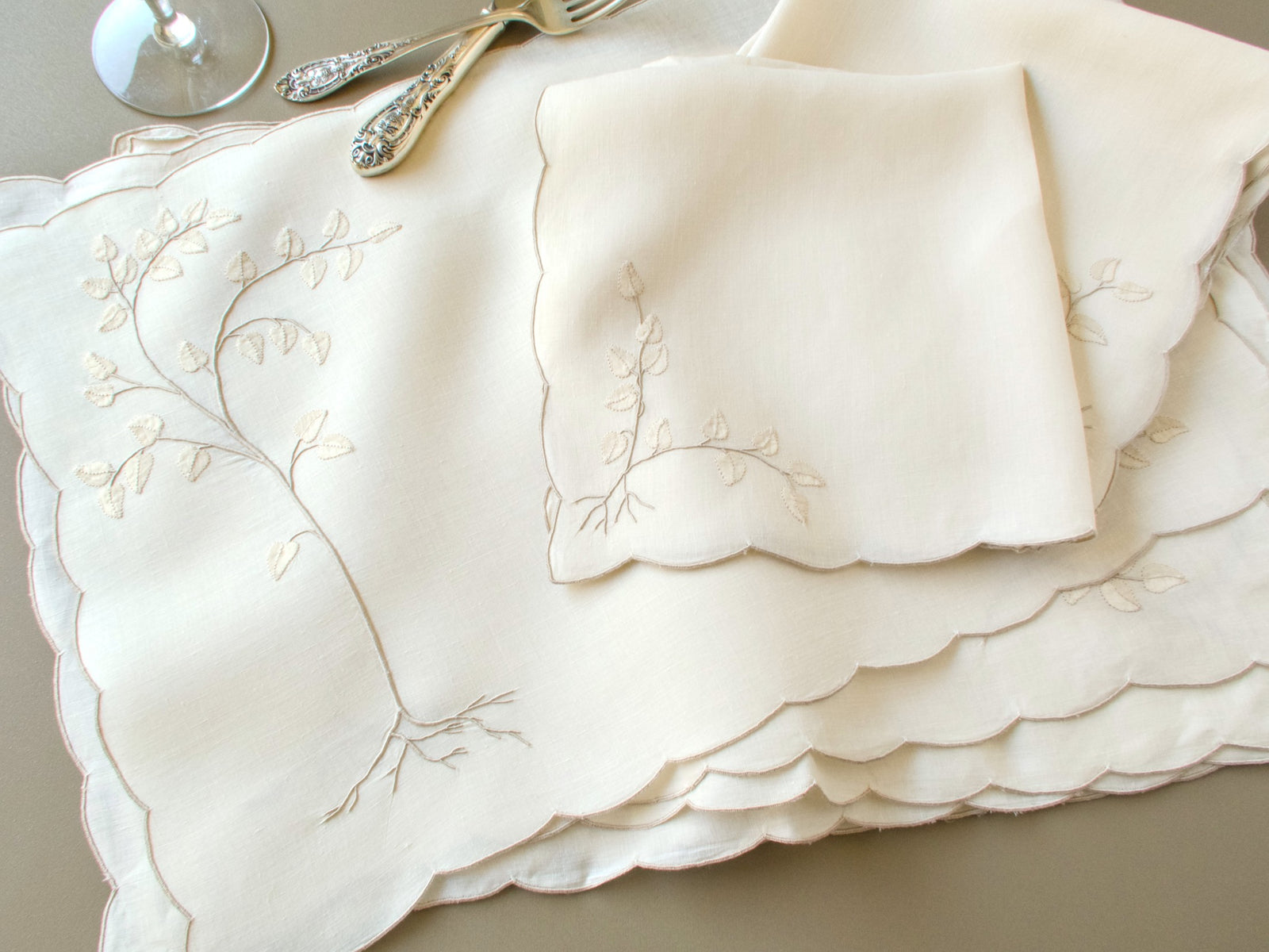 Stack of vintage Madeira linen placemats with scalloped edges and a matching napkin beside antique silver flatware