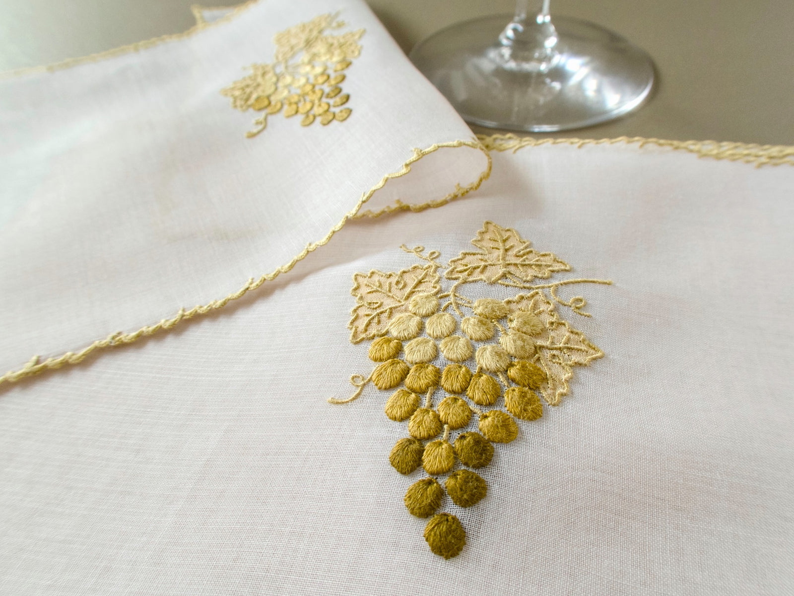 Close view of a vintage Marghab "Grape & Leaf" organdy cocktail napkin with a hand embroidered cluster of grapes shading from pale gold to olive, appliquéd leaves and a gold scalloped edge, beside a wine glass