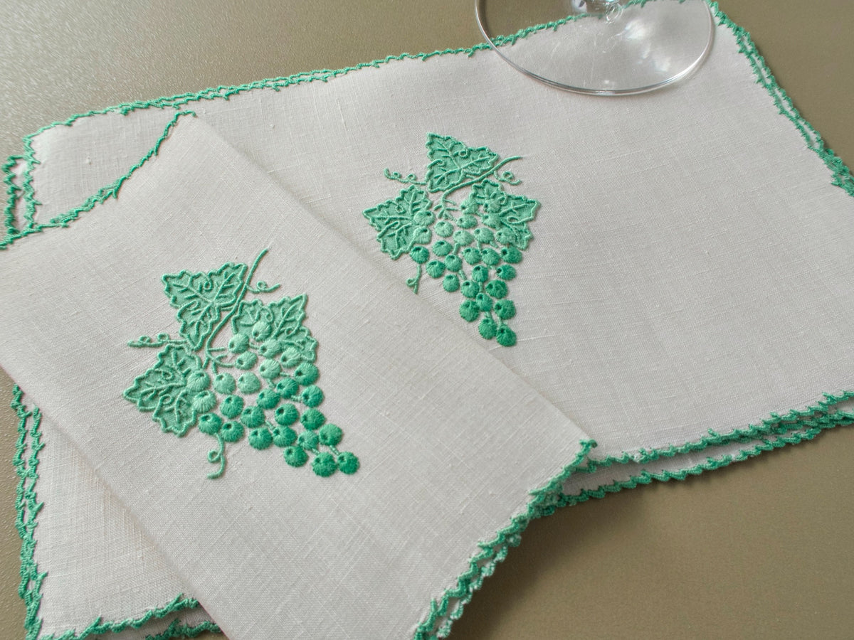 Two vintage Marghab "Grape &amp; Leaf" linen cocktail napkins hand embroidered in Madeira with green grape clusters and appliquéd leaves and a green scalloped edge, beside a wine glass