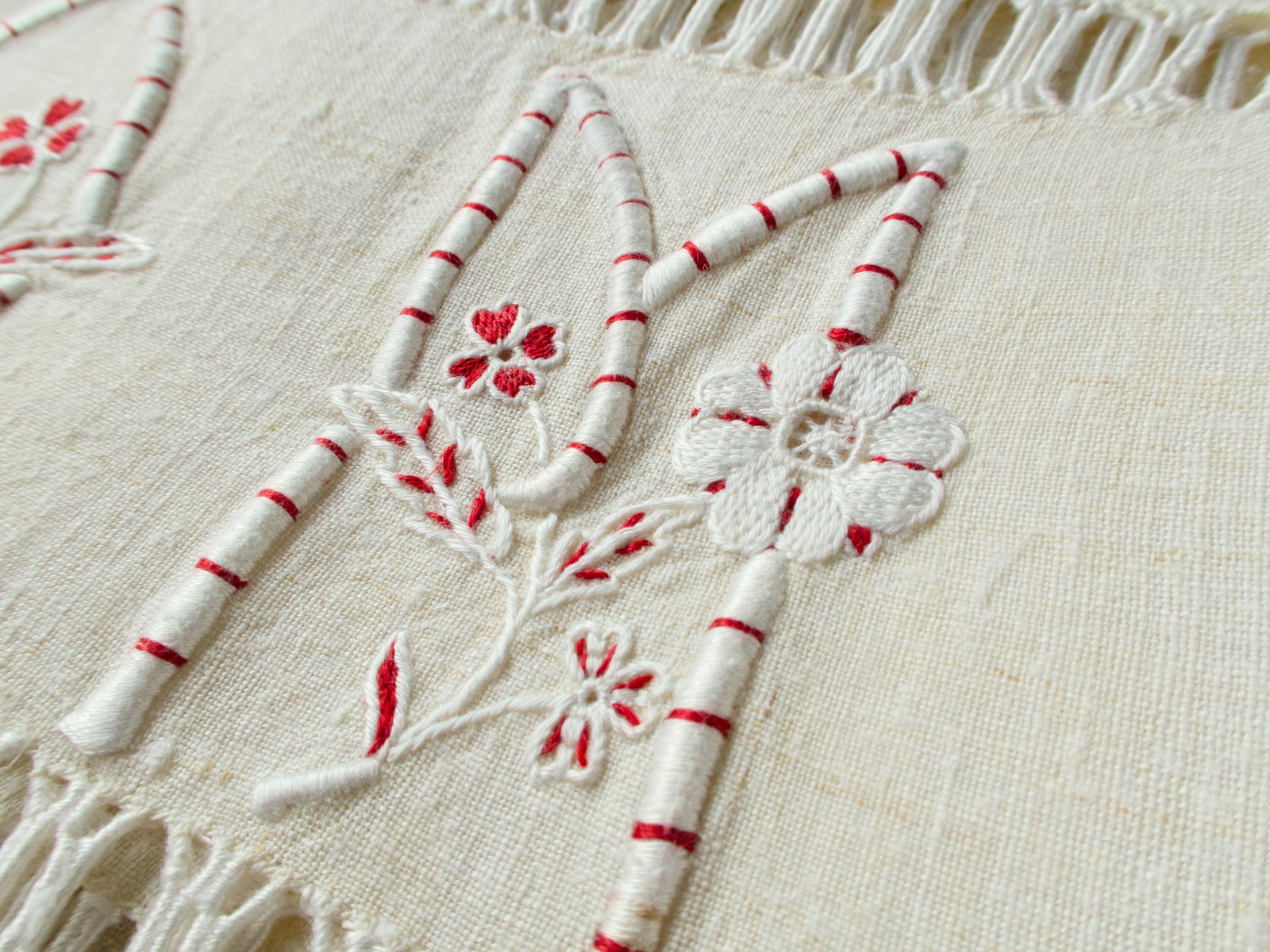 Close-up of vintage French homespun linen towel hand embroidered with a bold “OM” monogram in white padded satin stitch, red accent detailing and small flowers, with fringe