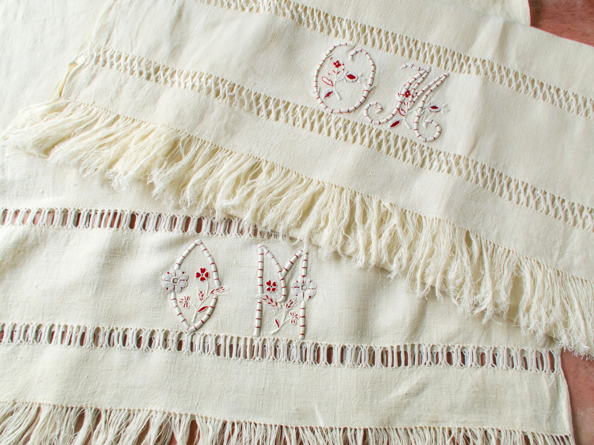 Two vintage French homespun linen towels side by side showing the slight variation between the two hand embroidered “OM” monograms with fringe