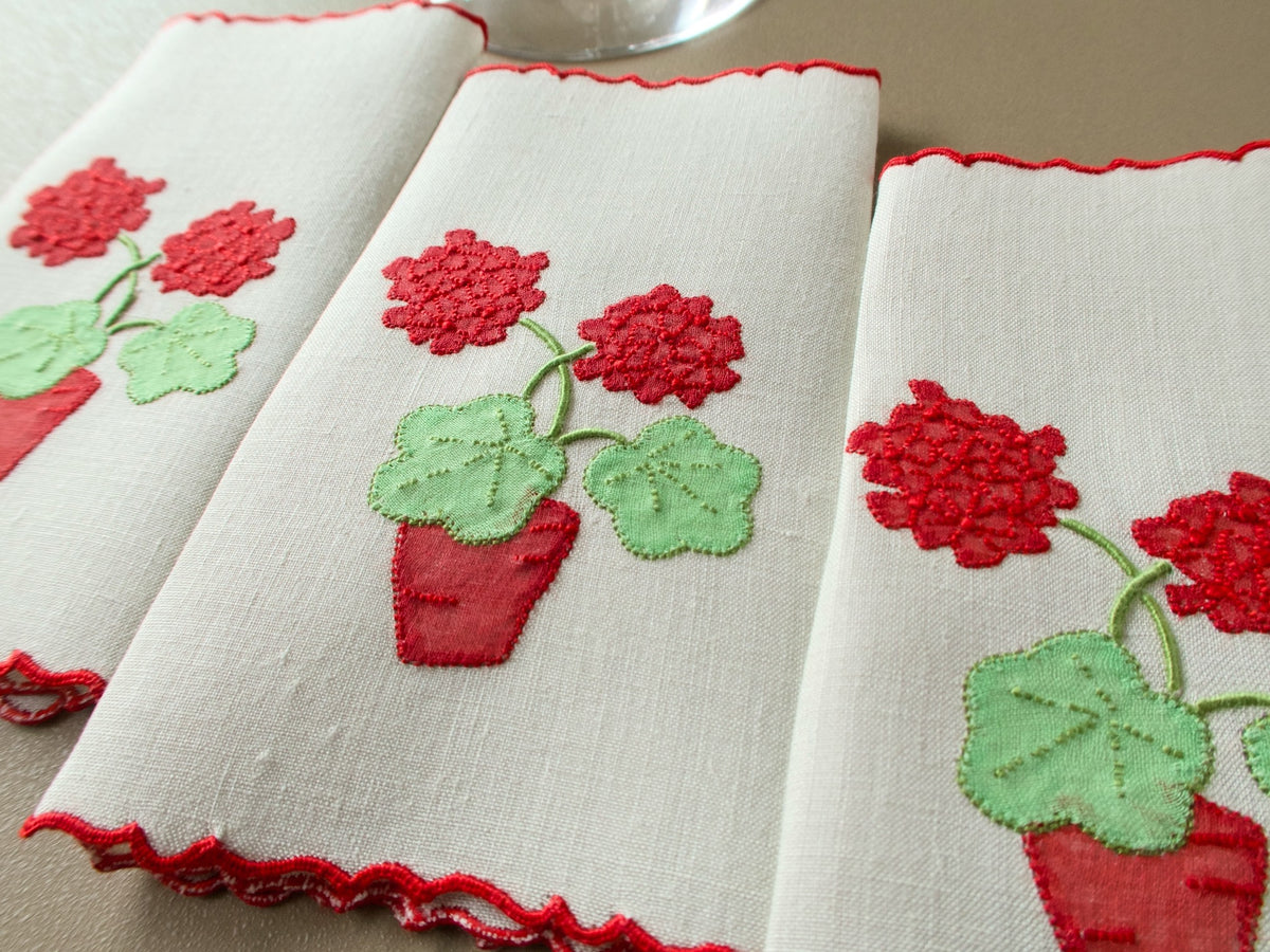 Three vintage Marghab "Geranium" cocktail napkins laid side by side, each with a pot of red geraniums and a red scalloped edge