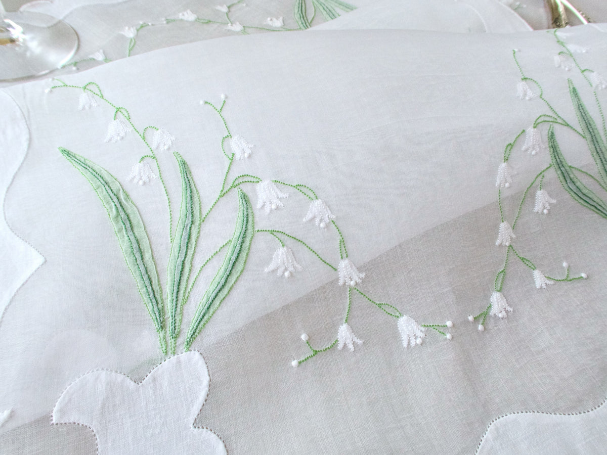 Close-up detail of hand embroidered lily of the valley on vintage Madeira organdy placemat with white blossoms and green leaves in shadow work.