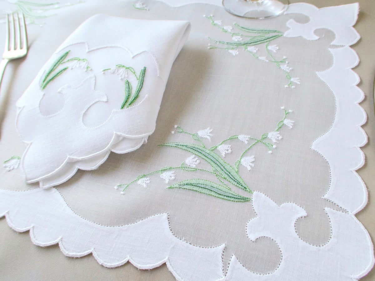 Vintage Madeira lily of the valley organdy placemat with scalloped cutwork border and folded linen napkin on beige background.