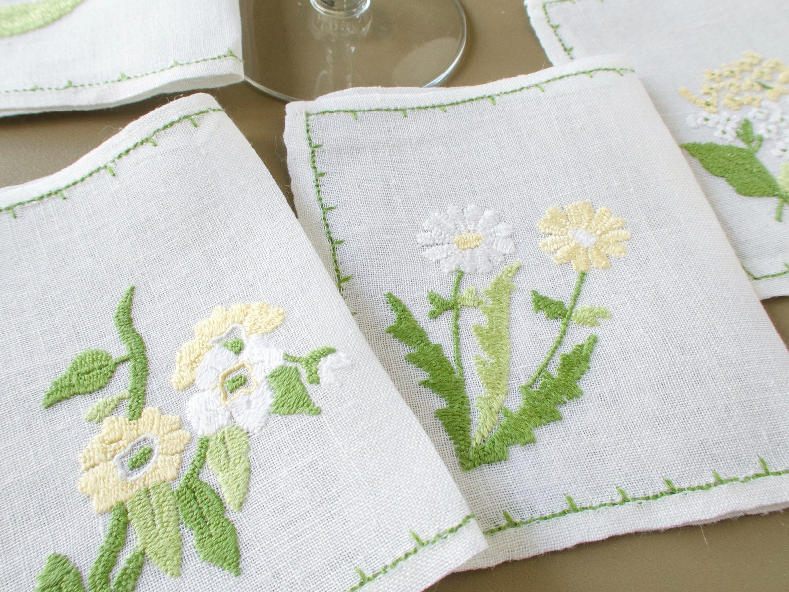 Close view of vintage Baroness Rapisardi floral cocktail napkins with soft yellow and white flowers, green foliage and hand-stitched borders, with wine glass 