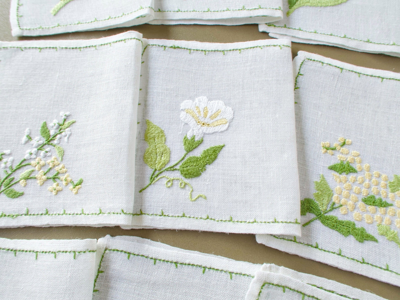 Three Baroness Rapisardi floral cocktail napkins with distinct botanical motifs and delicate green hand-stitched borders.