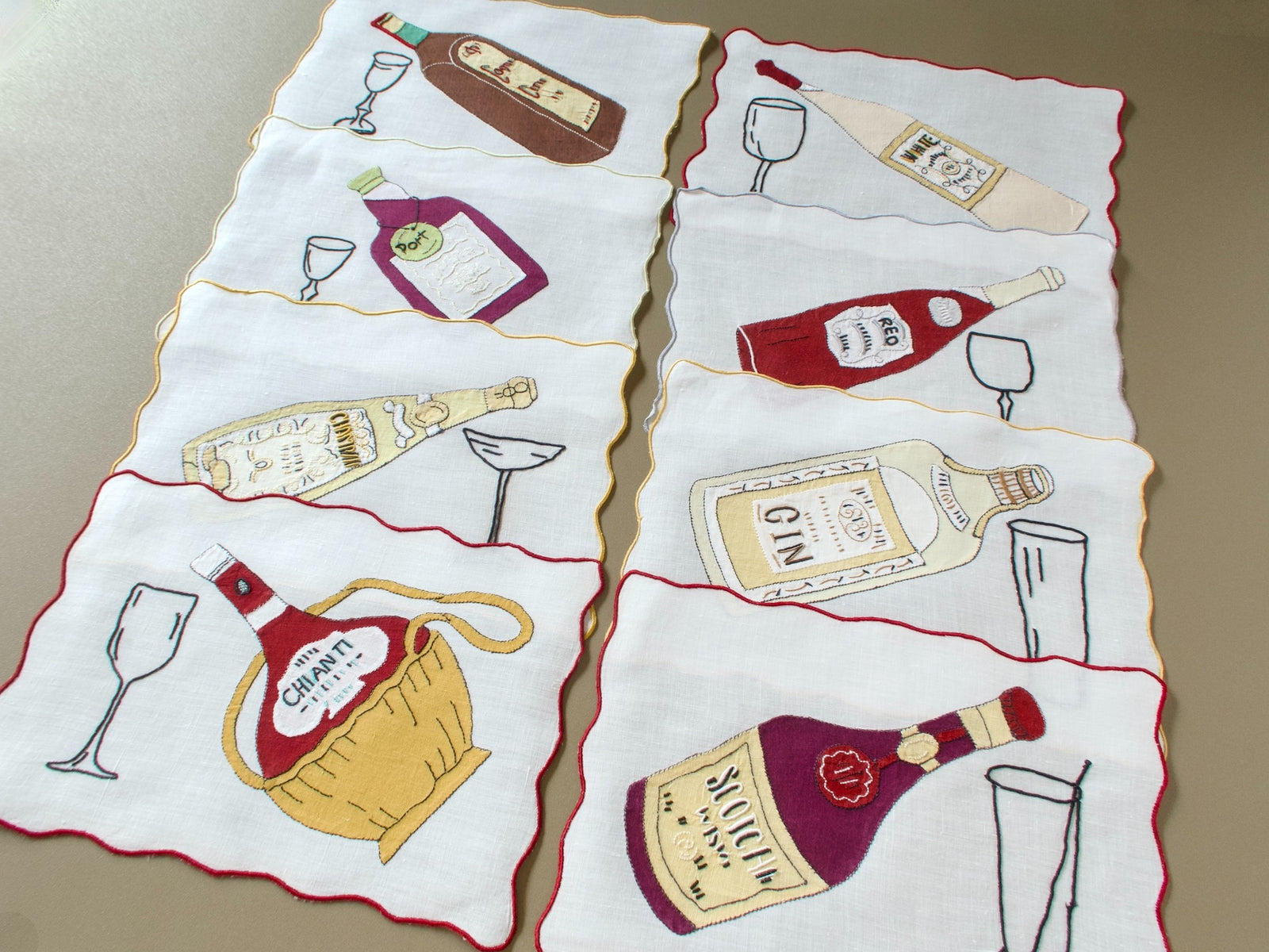 Eight vintage Madeira cocktail napkins laid out, each with a different appliqué bottle and embroidered glass, edged in red or gold
