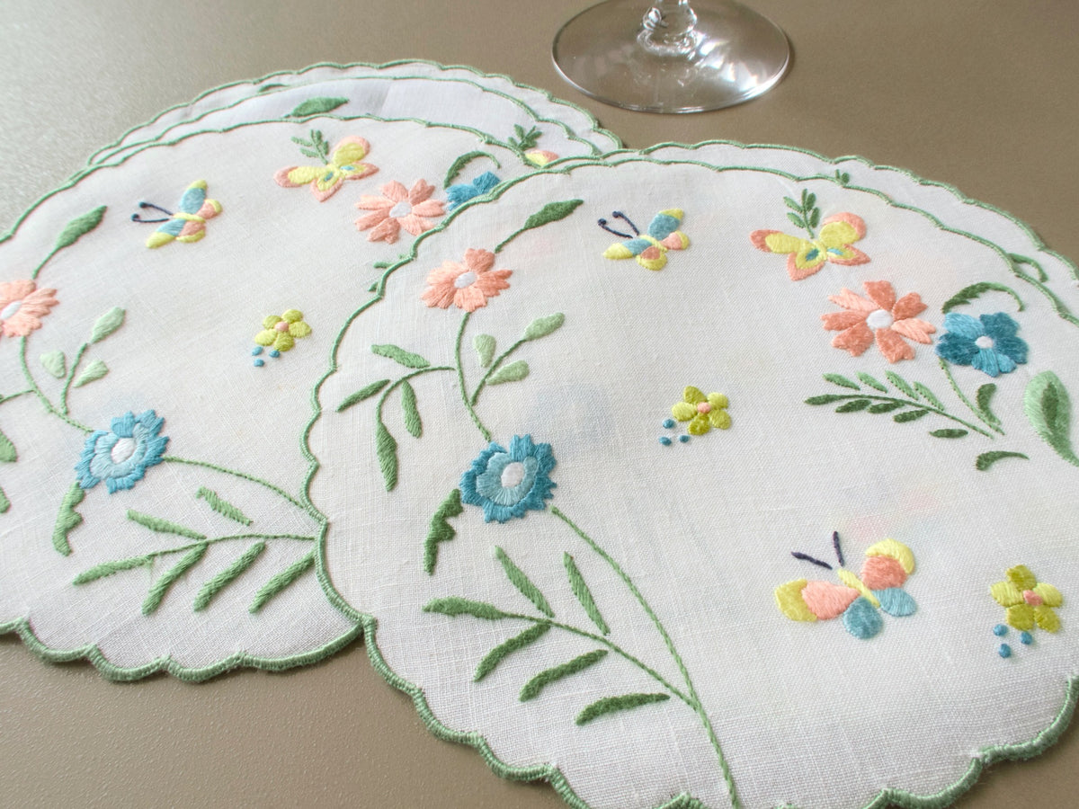 Two vintage Madeira linen cocktail rounds with embroidered butterflies and flowers and scalloped edges, beside a wine glass