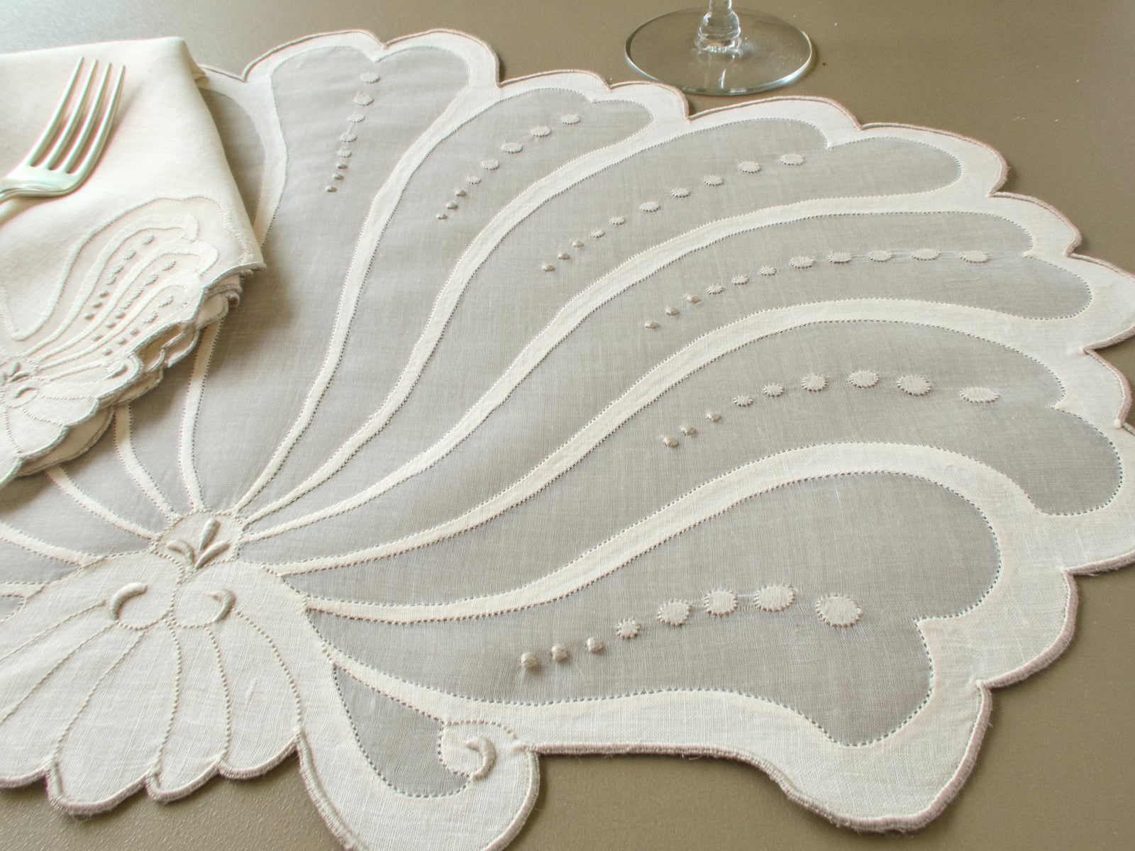 Close view of a vintage Madeira fan-shaped organdy and linen placemat with hand embroidered dots, beside a napkin and silver fork