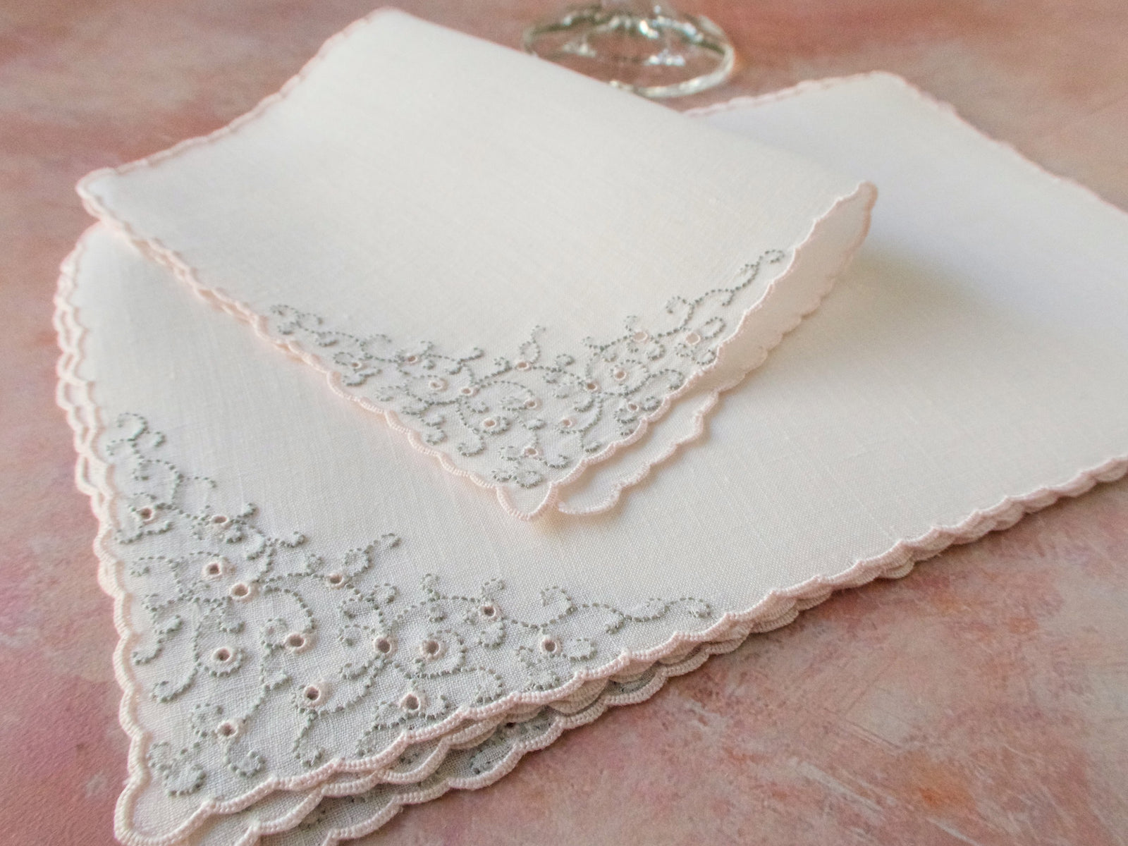 Folded vintage Marghab “Thousand Eye” cocktail napkin in pale pink Irish linen showing detailed Madeira eyelet embroidery along the corner.
