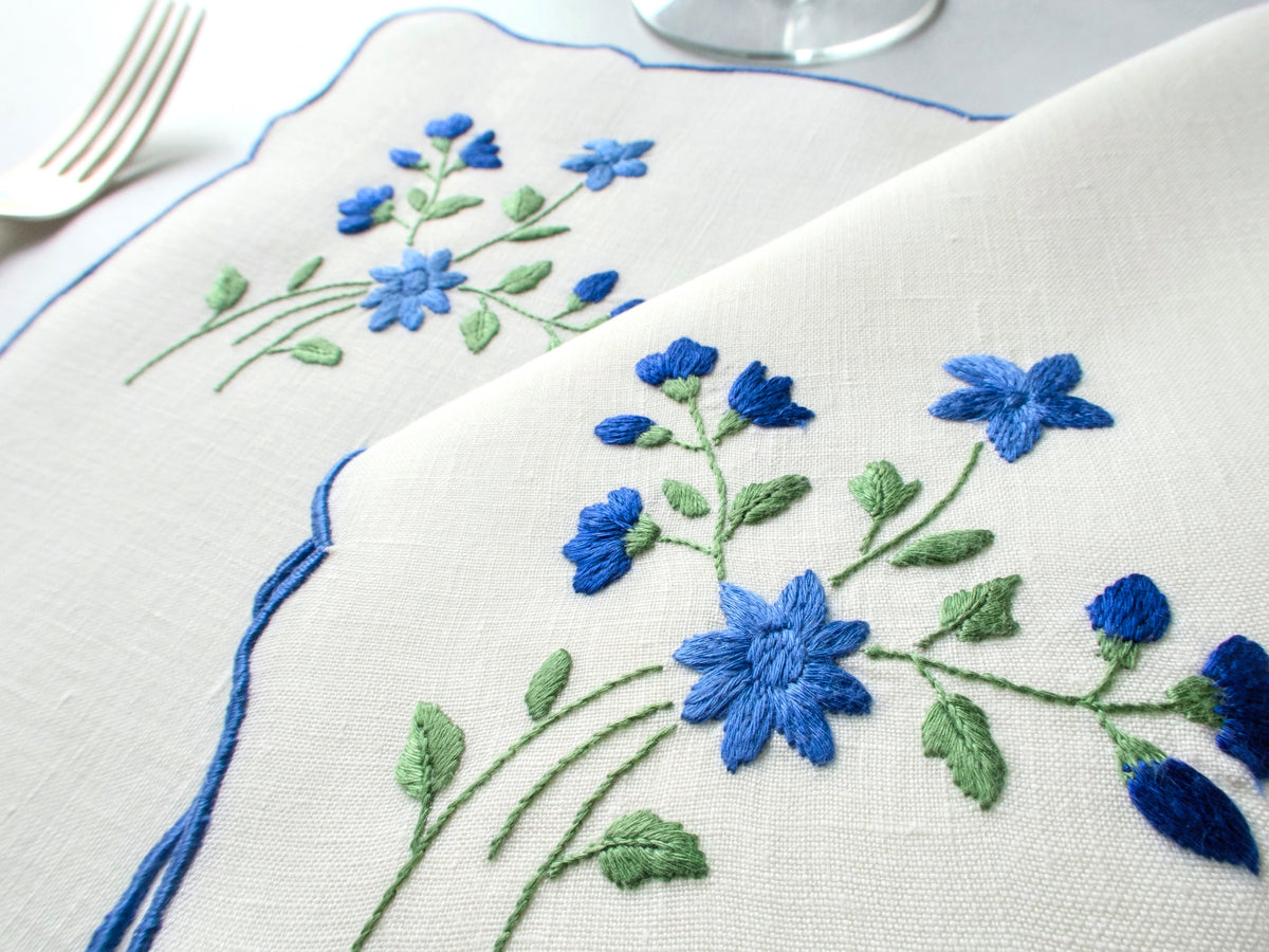 Vintage Madeira placemat close-up showing blue forget-me-not embroidery clusters with green leaves on off-white linen with blue scalloped border