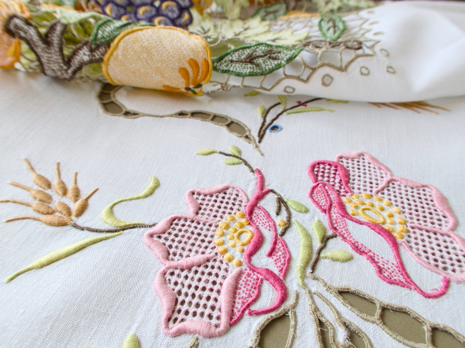 Close view of hand embroidered pink blossoms with openwork centers, wheat and a cutwork ribbon on a vintage Madeira tablecloth