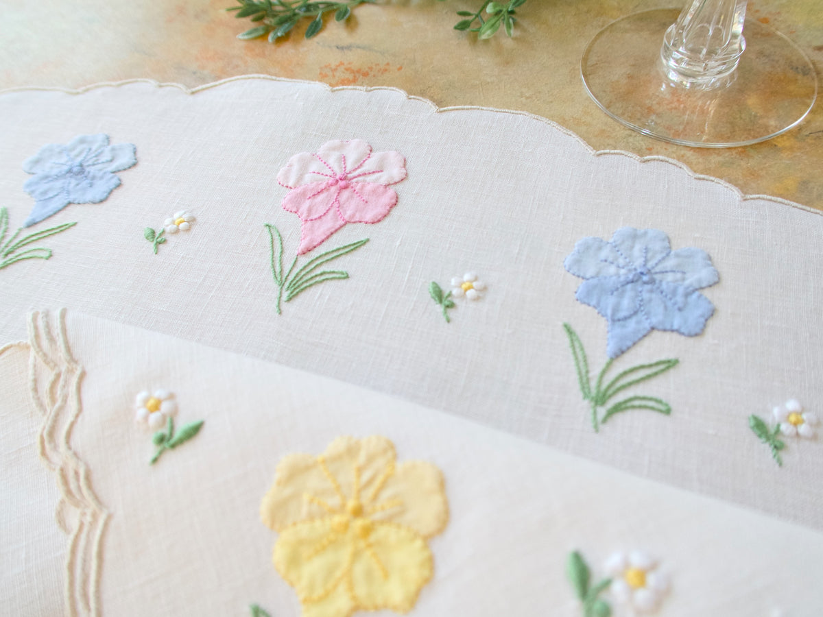 Close-up of vintage Madeira oval placemat on natural linen showing hand embroidered spring flowers in pink, blue, and yellow with scalloped edges.