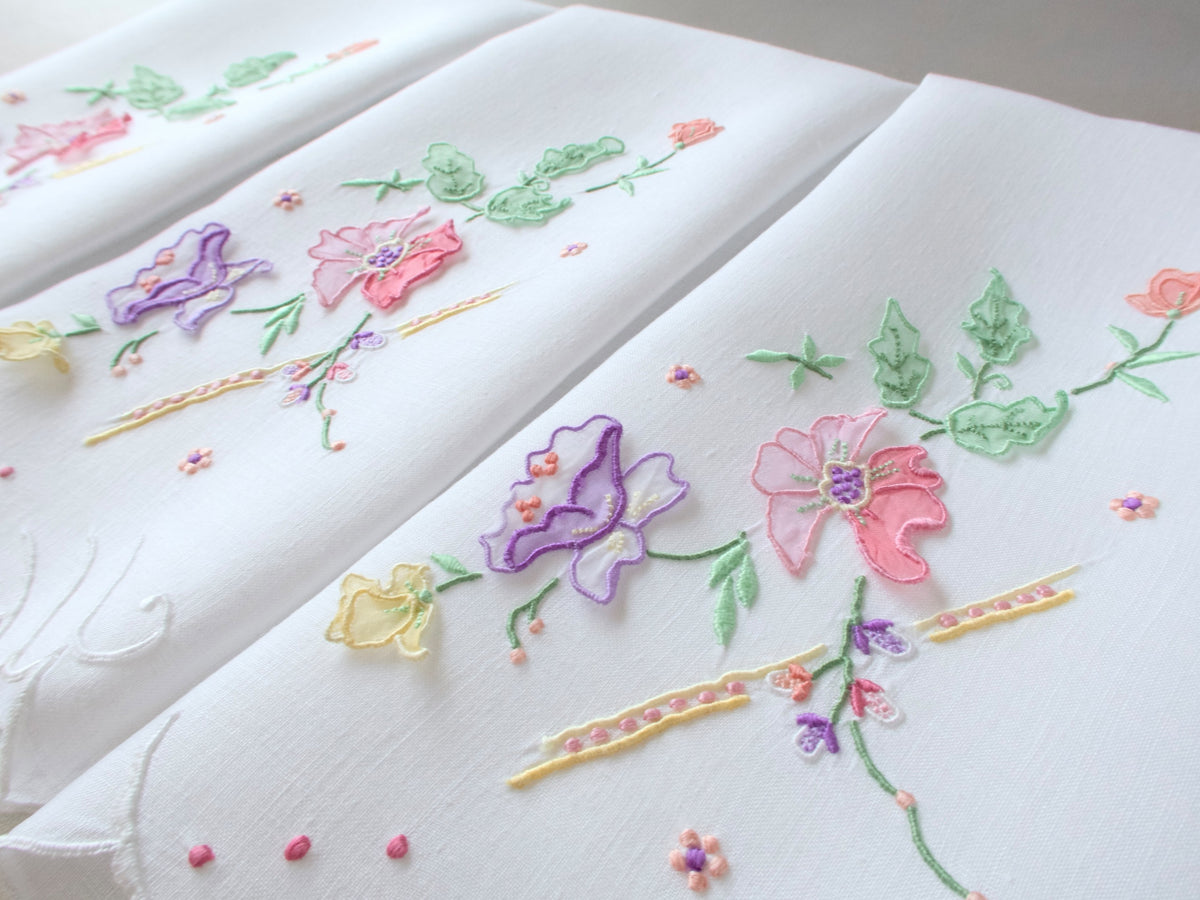 Three vintage Madeira guest towels fanned out showing colorful trembler flower embroidery in pastel pink, purple, and yellow.