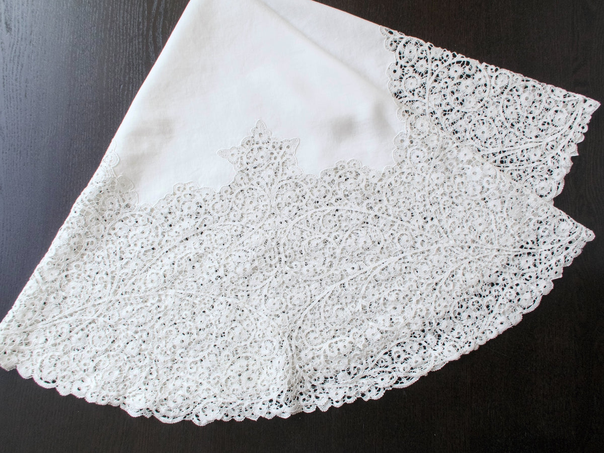 Round Italian Cantù lace tablecloth draped against a dark background, showing its linen center and wide lace border.