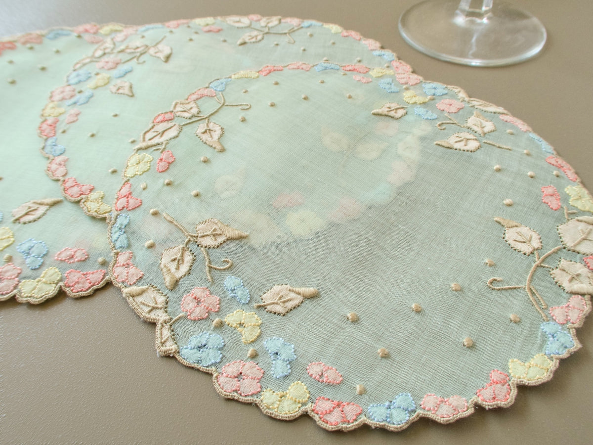 Vintage Madeira organdy cocktail rounds on beige background, pale green sheer organdy with multicolor pink blue and yellow flower border and embroidered vines with granitos.
