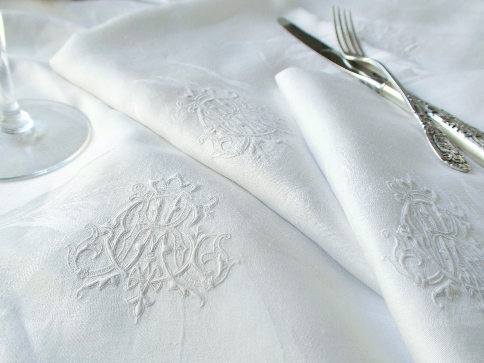 Antique damask linen napkin set on a table beside a wine glass and silver flatware, showing the embroidered ACR monogram