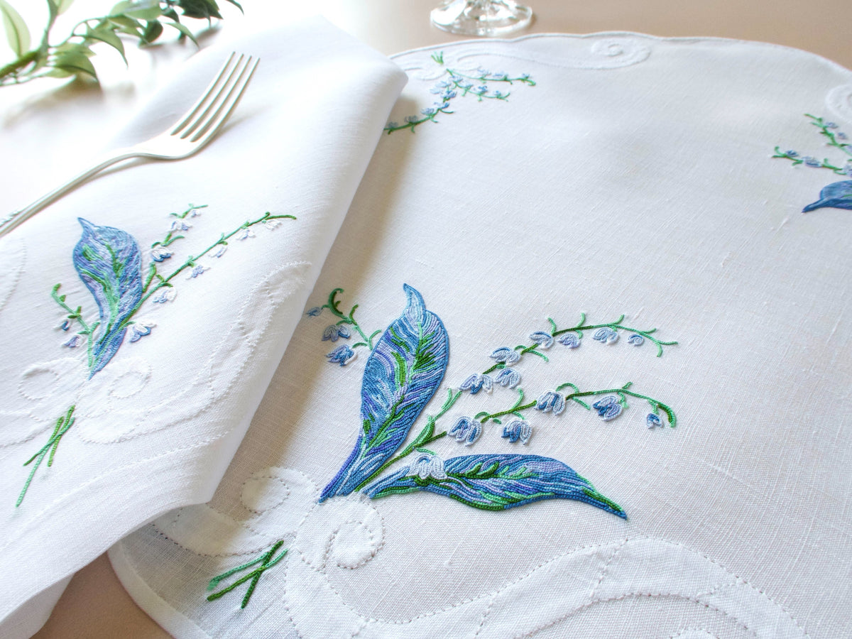 Vintage D. Porthault Lily of the Valley placemat with folded linen napkin showing Point de Beauvais embroidery in rich blue and green on white linen.